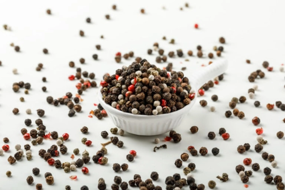 Un petit bol blanc rempli de grains de poivre noir et rouge séchés, entouré de grains dispersés sur fond blanc.